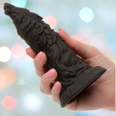 A hand with pink-painted nails holds the Monster Dong The Werewolf by CalExotics, a black sculpted toy shaped like a wolf’s head and fur with a broad base and textured details, set against a softly blurred, colorful background.